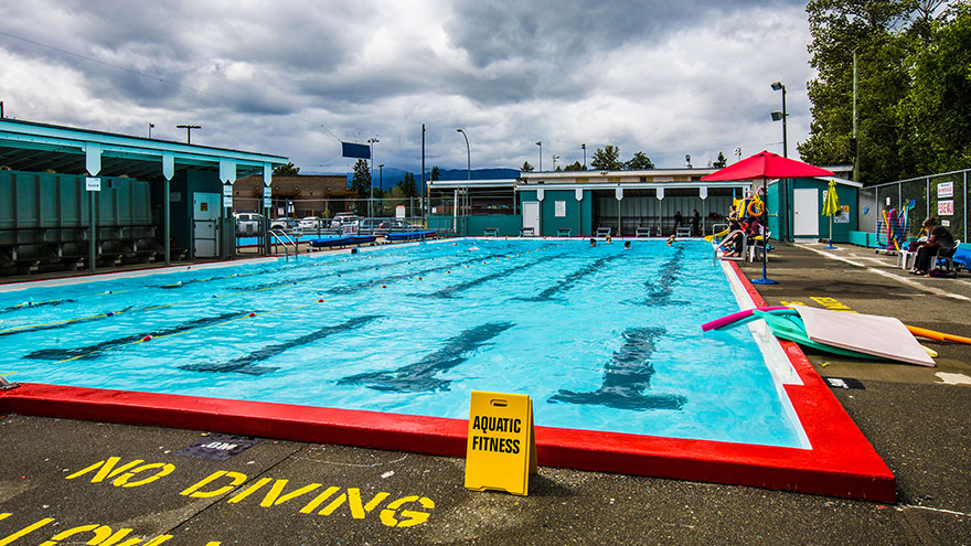 A large outdoor public pool with several people in the distance taking an aquafit class. The water is turquoise and the sky is cloudy.