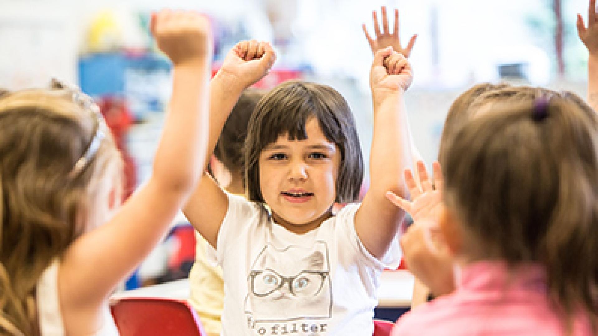 A young girl in a classroom enthusiastically raises both hands while smiling, surrounded by other children.