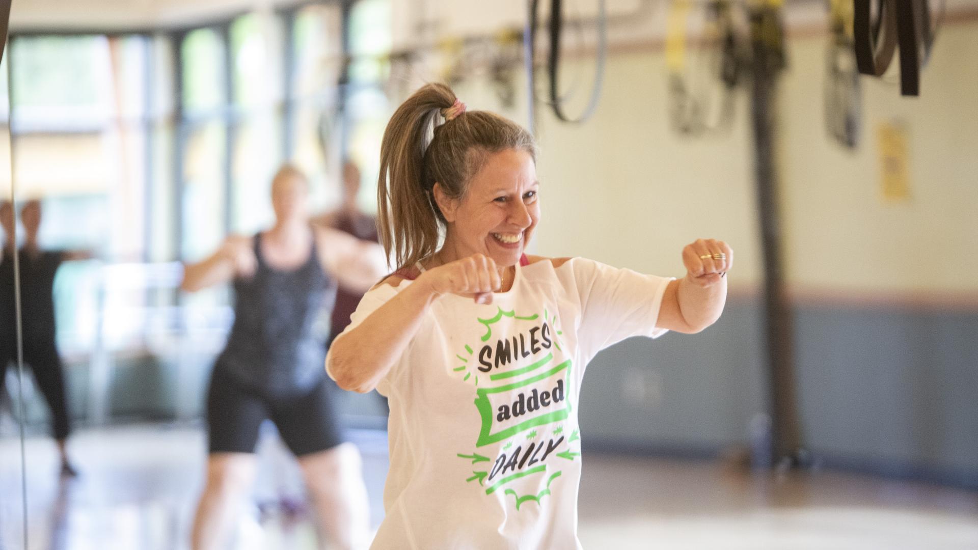 A smiling woman participating in a fitness class, her shirt says "smiles added daily"