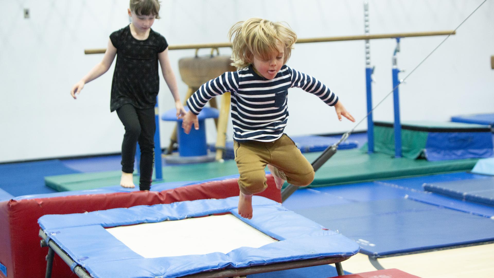 Two children participating in a gymnastics class. One is stepping onto a small trampoline, while the other is mid-air, jumping from that trampoline onto a mat.