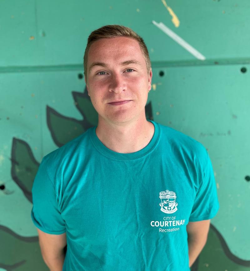 A man in a teal "City of Courtenay Recreation" shirt stands in front of a green mural background, smiling slightly.