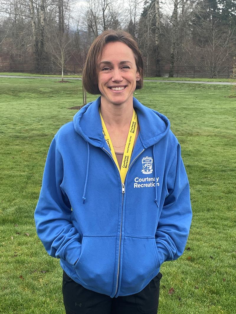 A woman in a blue "Courtenay Recreation" hoodie stands on a grassy field with bare trees in the background, smiling warmly.