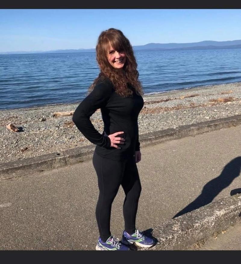 A woman in black activewear smiles while standing on a paved walkway beside a rocky beach and calm ocean.