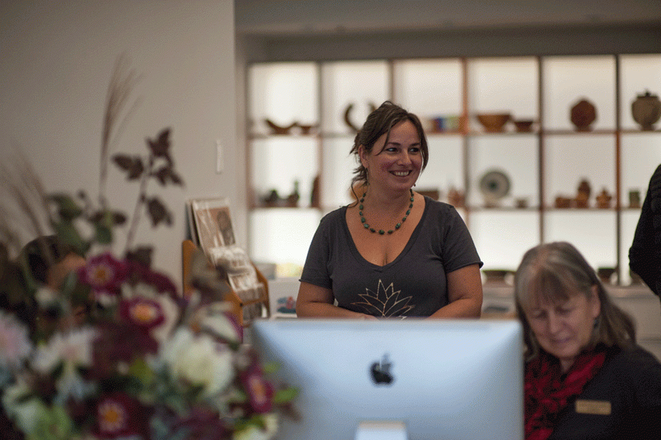 A woman standing behind a front desk smiling, with another woman seated in front of an iMac computer; shelves with artwork and pottery in the background.