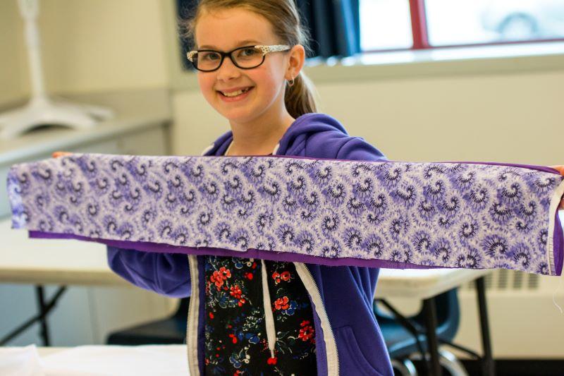 A smiling girl holding up a long, purple-patterned fabric piece in a classroom setting, displaying her sewing project.
